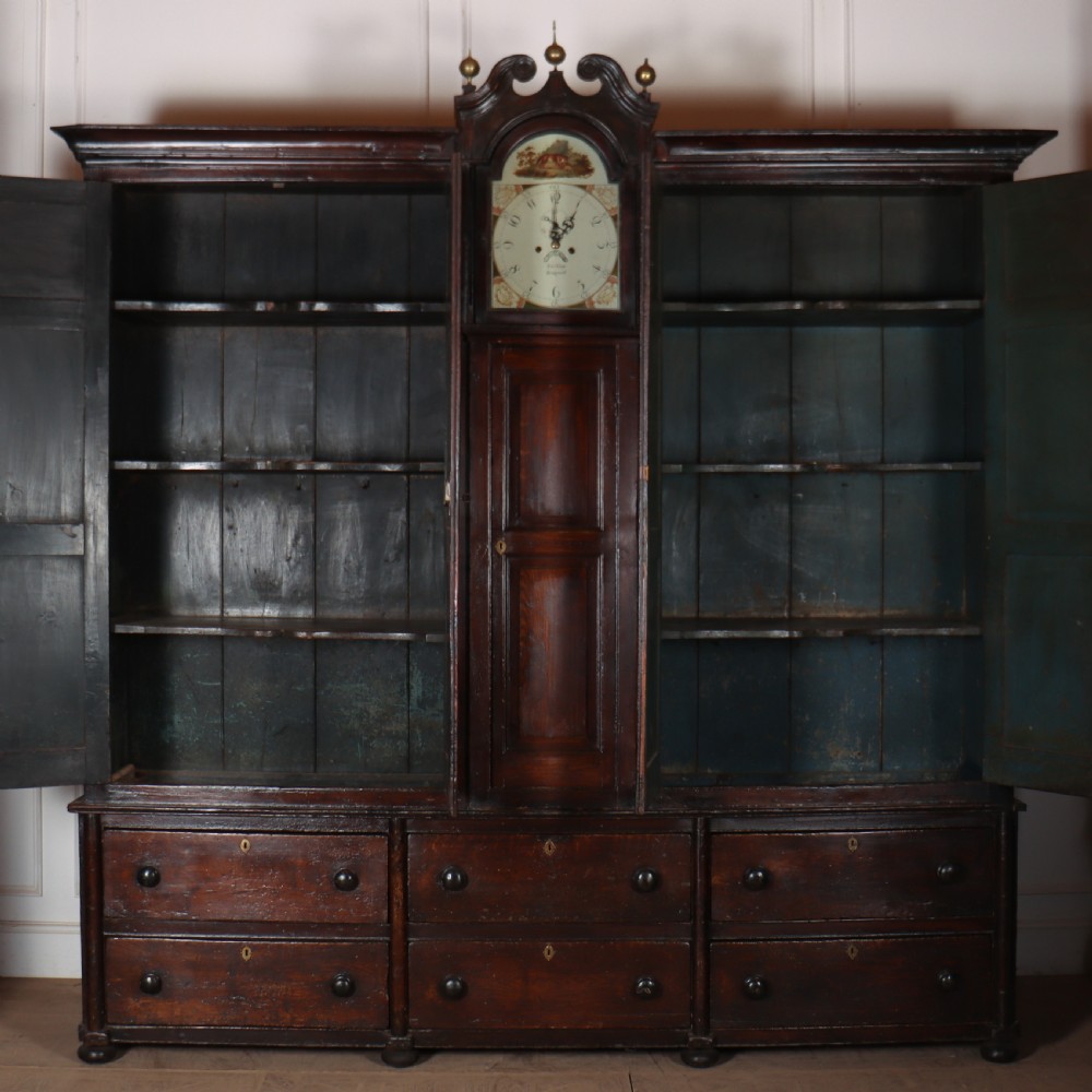 An antique dark brown wooden housekeepers cupboard featuring panelled doors, a central integrated grandfather clock, and a base section with six drawers. View IX