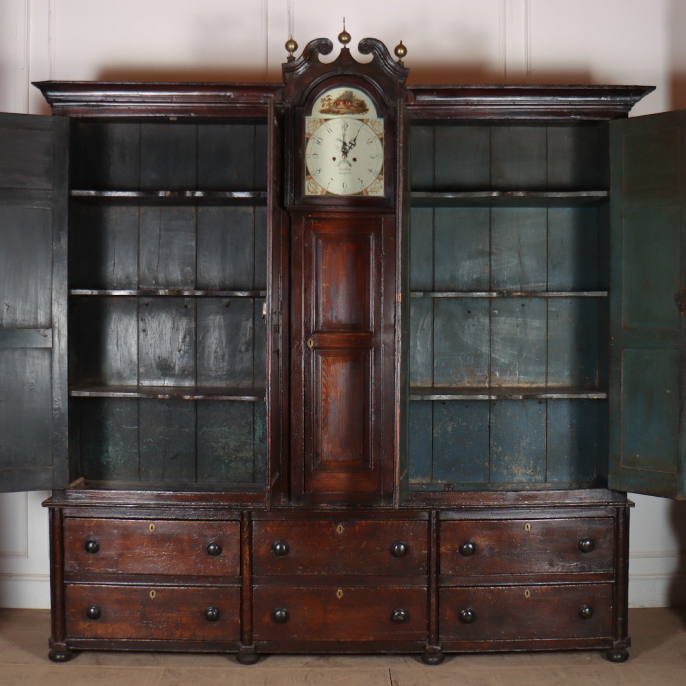 An antique dark brown wooden housekeepers cupboard featuring panelled doors, a central integrated grandfather clock, and a base section with six drawers. View X