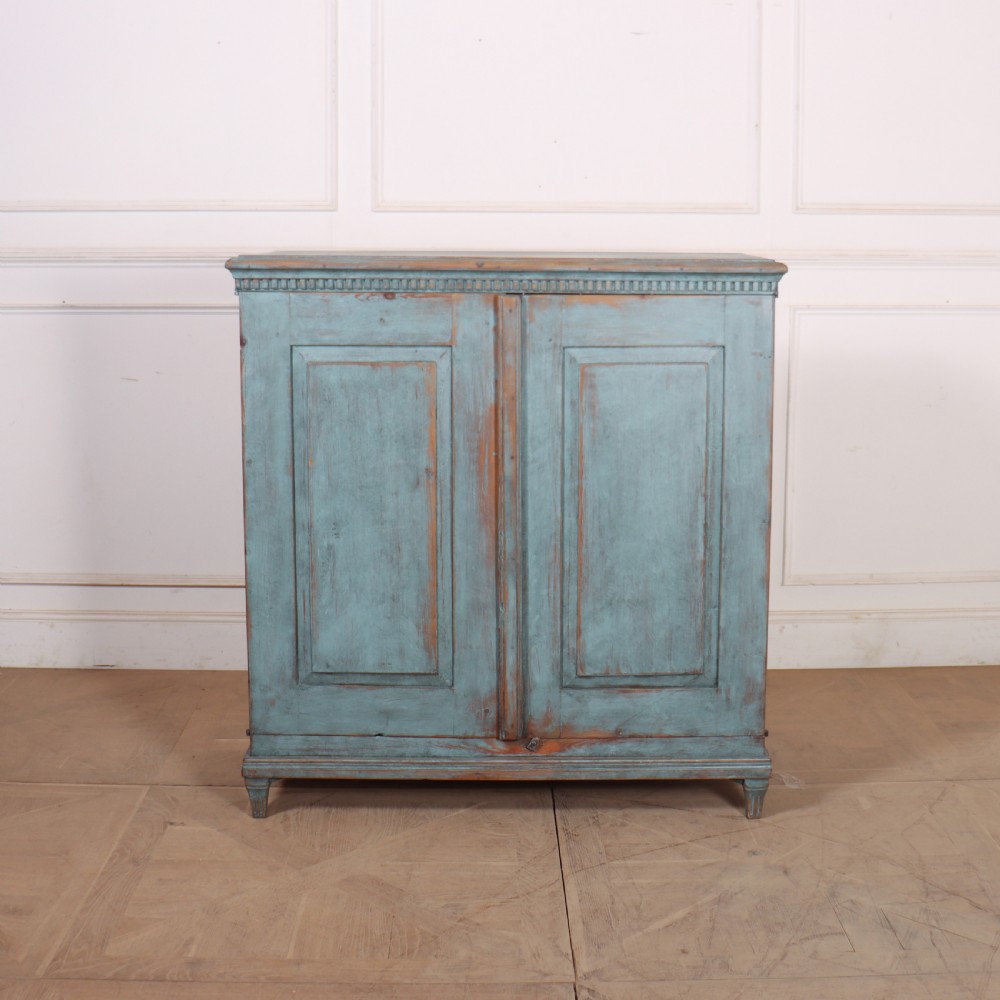 A Swedish two-door wooden cupboard featuring a distressed blue painted finish, panelled doors, dentil moulding along the top frieze, and short tapered legs.
