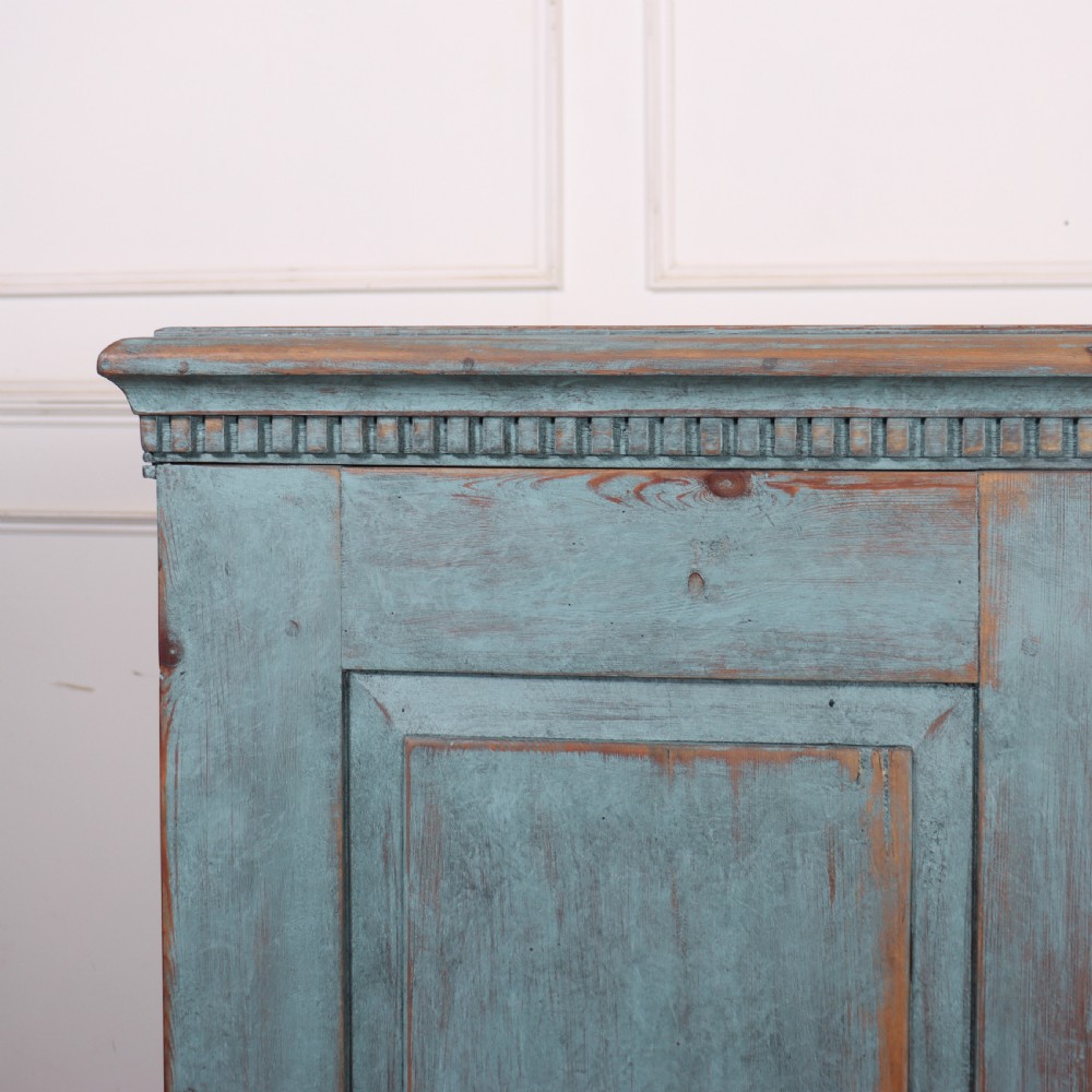 A Swedish two-door wooden cupboard featuring a distressed blue painted finish, panelled doors, dentil moulding along the top frieze, and short tapered legs. View II