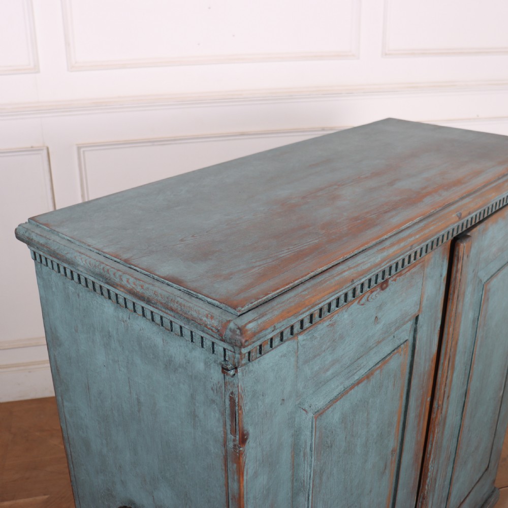 A Swedish two-door wooden cupboard featuring a distressed blue painted finish, panelled doors, dentil moulding along the top frieze, and short tapered legs. View X