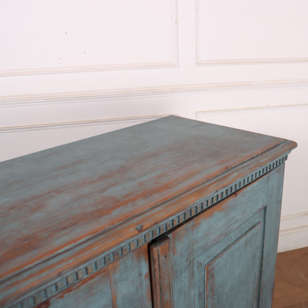 A Swedish two-door wooden cupboard featuring a distressed blue painted finish, panelled doors, dentil moulding along the top frieze, and short tapered legs. View XI