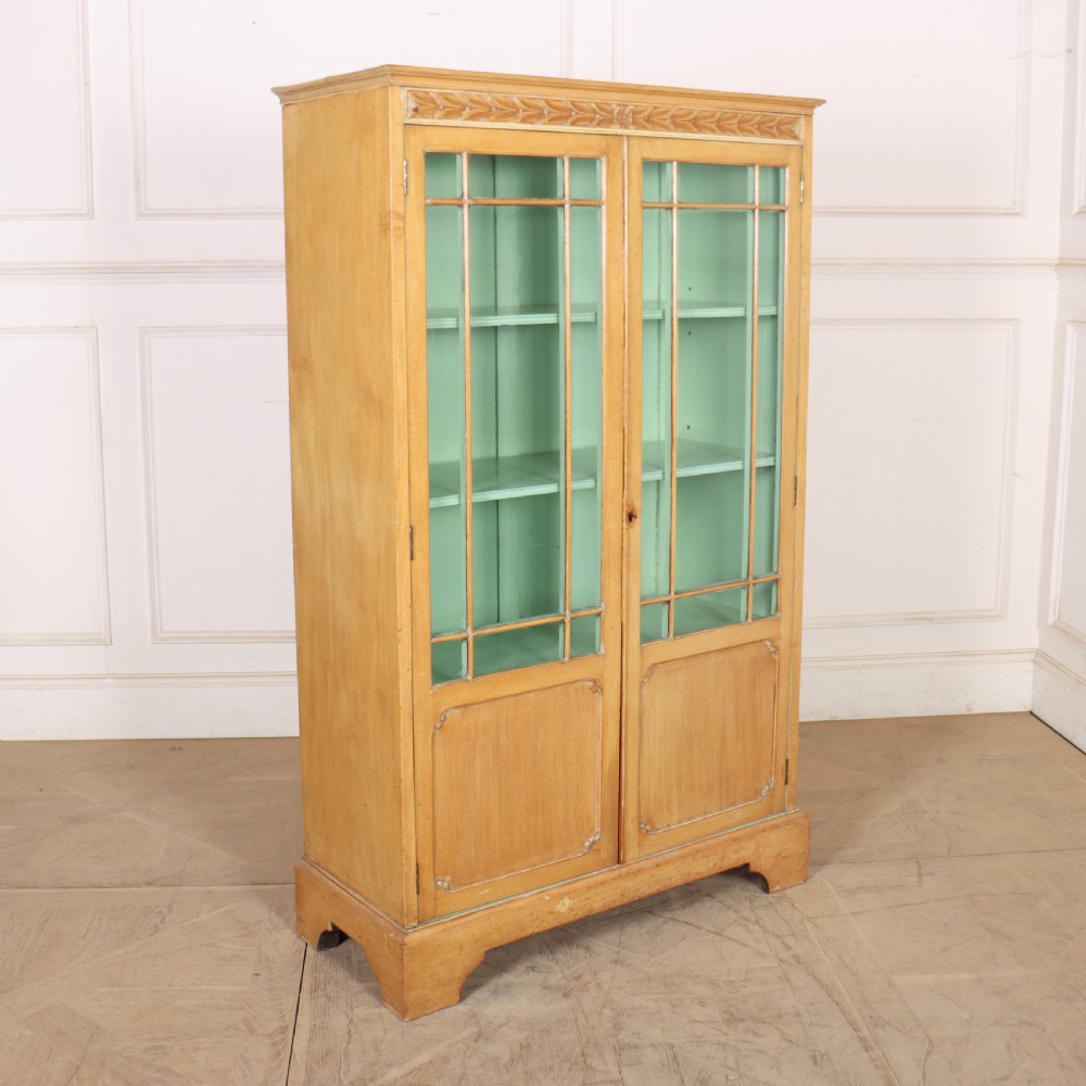 A tall English bleached walnut bookcase featuring two glazed doors with geometric glazing bars, a carved leaf frieze at the top, and a contrasting mint green painted interior.