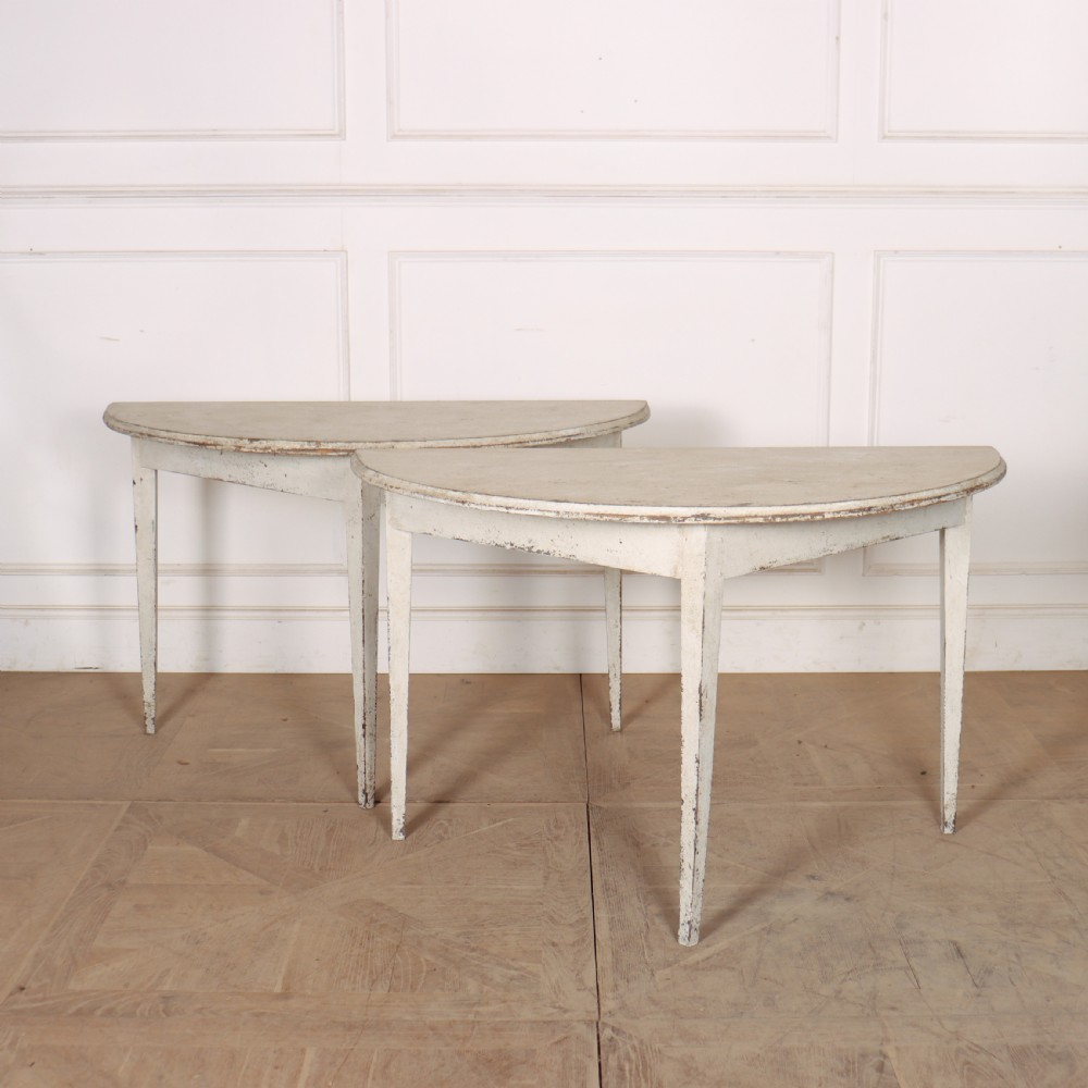 A pair of Swedish demi-lune console tables with distressed off-white painted finish and tapered legs.