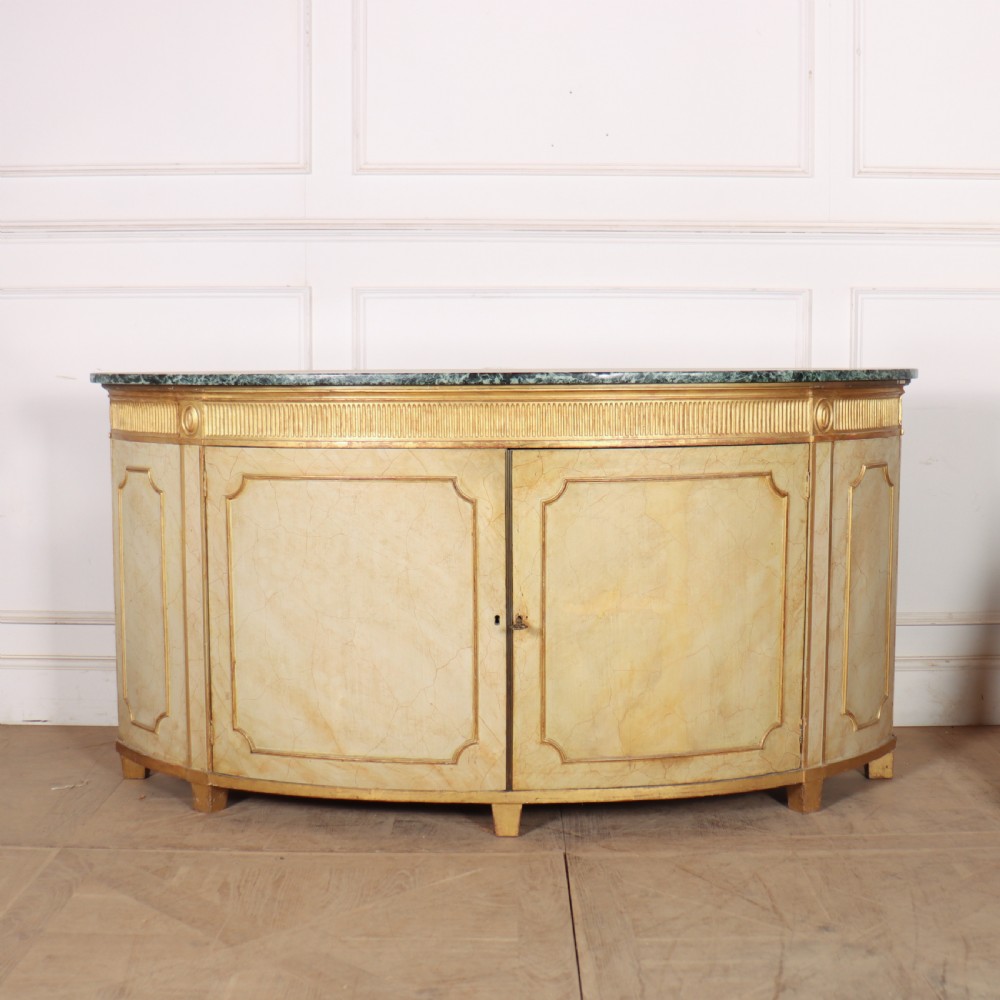 A Neoclassical style cream painted demi-lune side cabinet featuring a dark green marble top, fluted gilded frieze, and panelled cupboard doors.