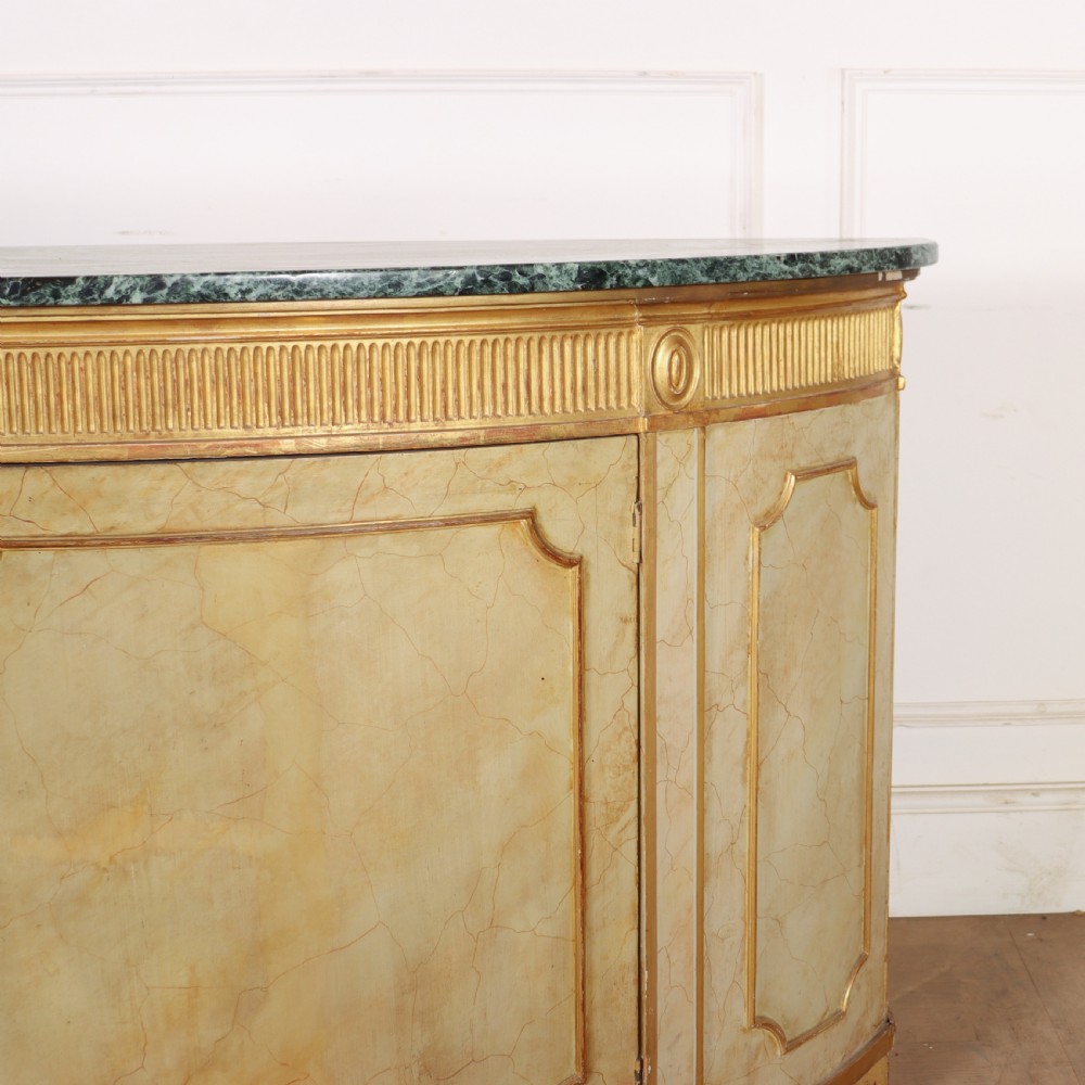 A Neoclassical style cream painted demi-lune side cabinet featuring a dark green marble top, fluted gilded frieze, and panelled cupboard doors. View V