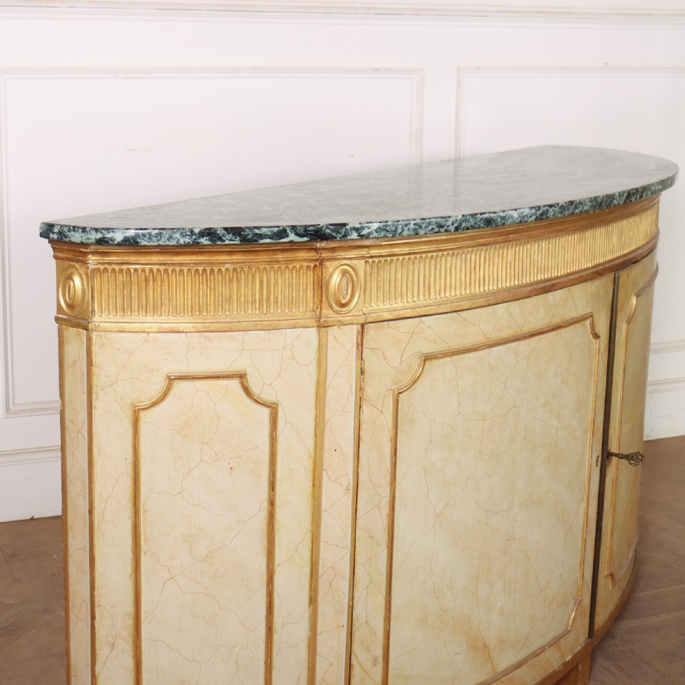 A Neoclassical style cream painted demi-lune side cabinet featuring a dark green marble top, fluted gilded frieze, and panelled cupboard doors. View IX