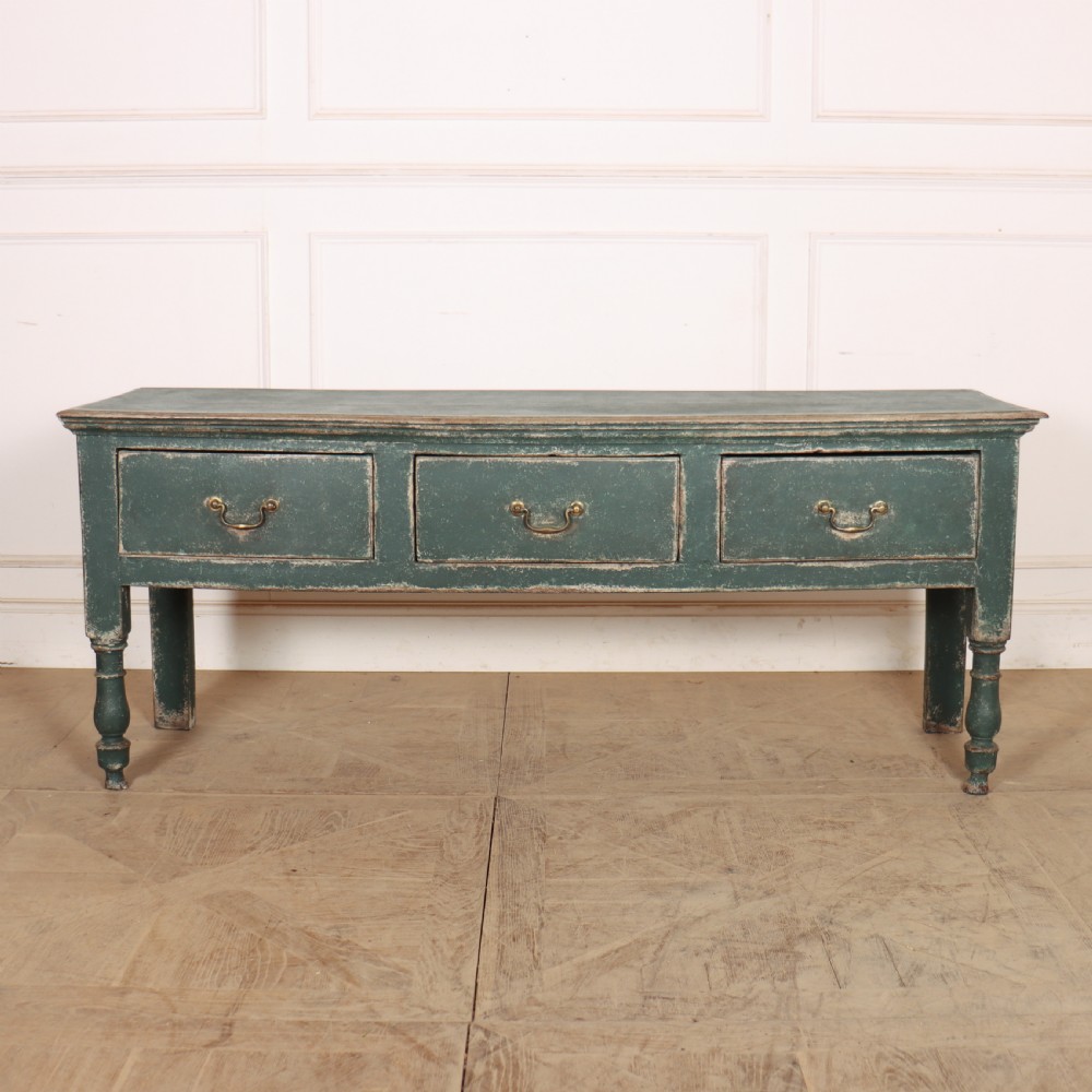 An English wooden dresser base painted in distressed muted green with three drawers and brass swan-neck handles.