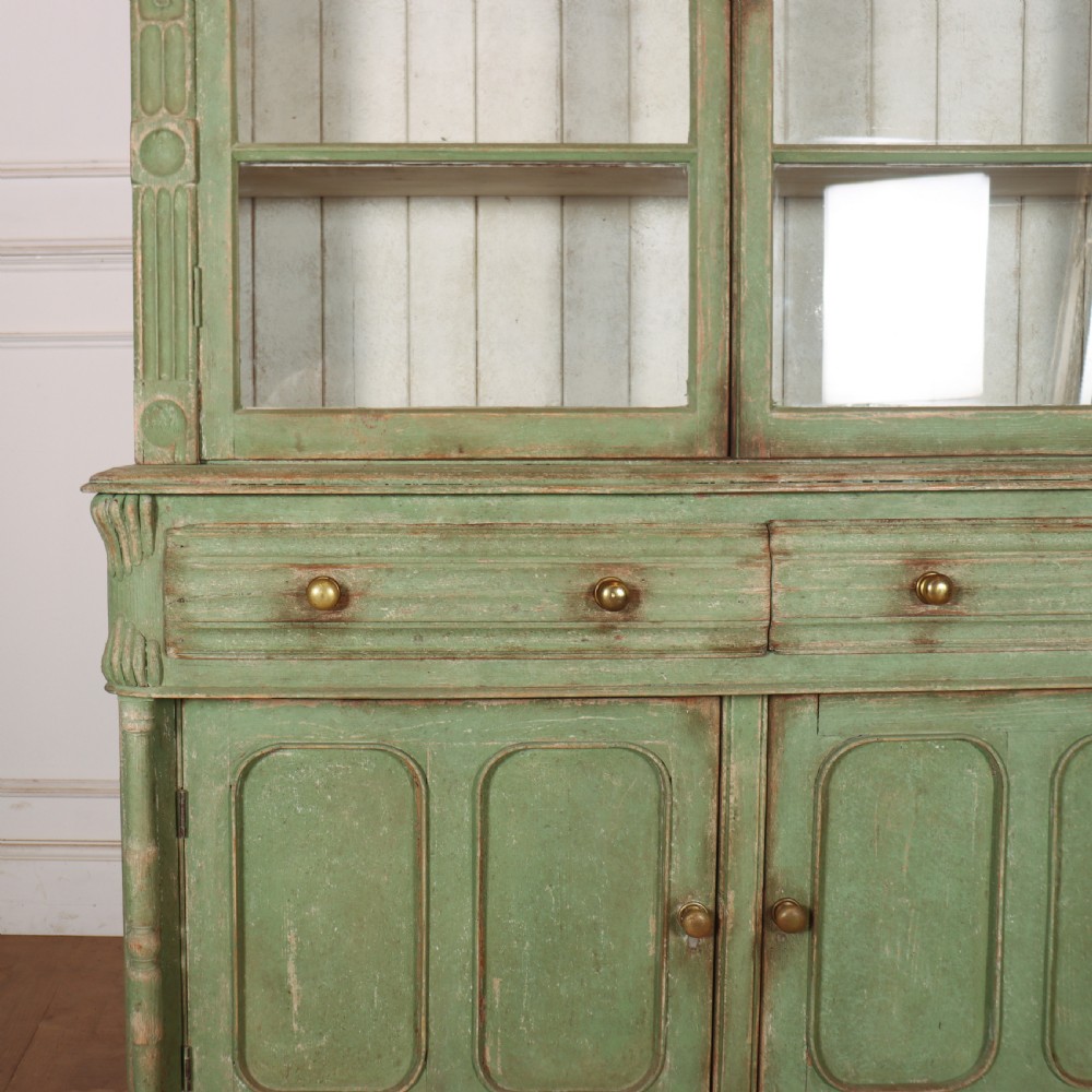 An antique West Country kitchen dresser in distressed sage green paint, featuring upper glazed doors and lower panelled cupboards. View VII