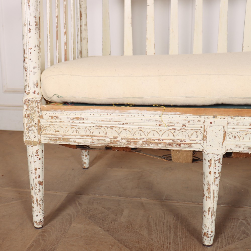 A long Gustavian style Swedish painted wooden settle bench featuring spindle sides and back, a distressed off-white finish, carved frame detailing, six tapered legs, and a fitted neutral upholstered cushion. View II