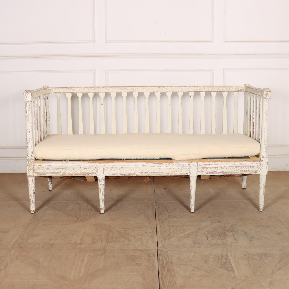 A long Gustavian style Swedish painted wooden settle bench featuring spindle sides and back, a distressed off-white finish, carved frame detailing, six tapered legs, and a fitted neutral upholstered cushion.