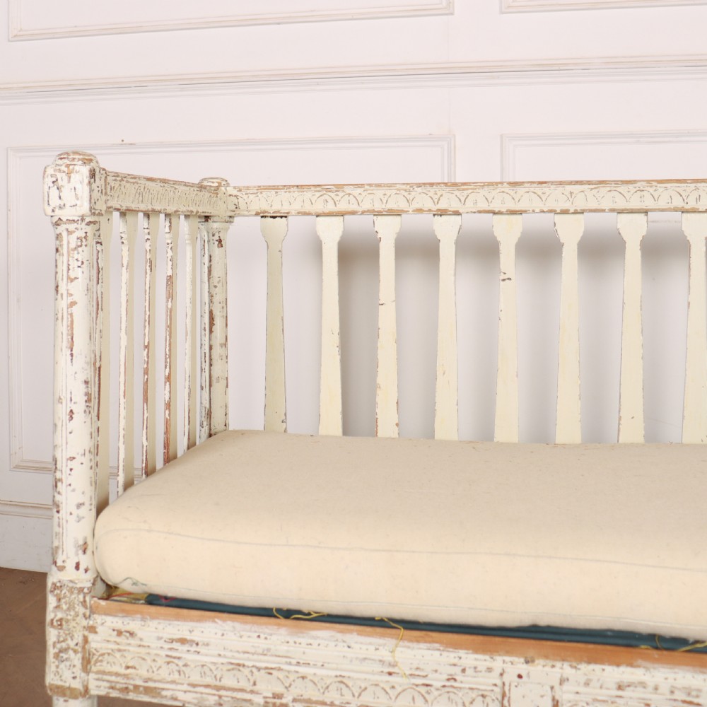 A long Gustavian style Swedish painted wooden settle bench featuring spindle sides and back, a distressed off-white finish, carved frame detailing, six tapered legs, and a fitted neutral upholstered cushion. View III
