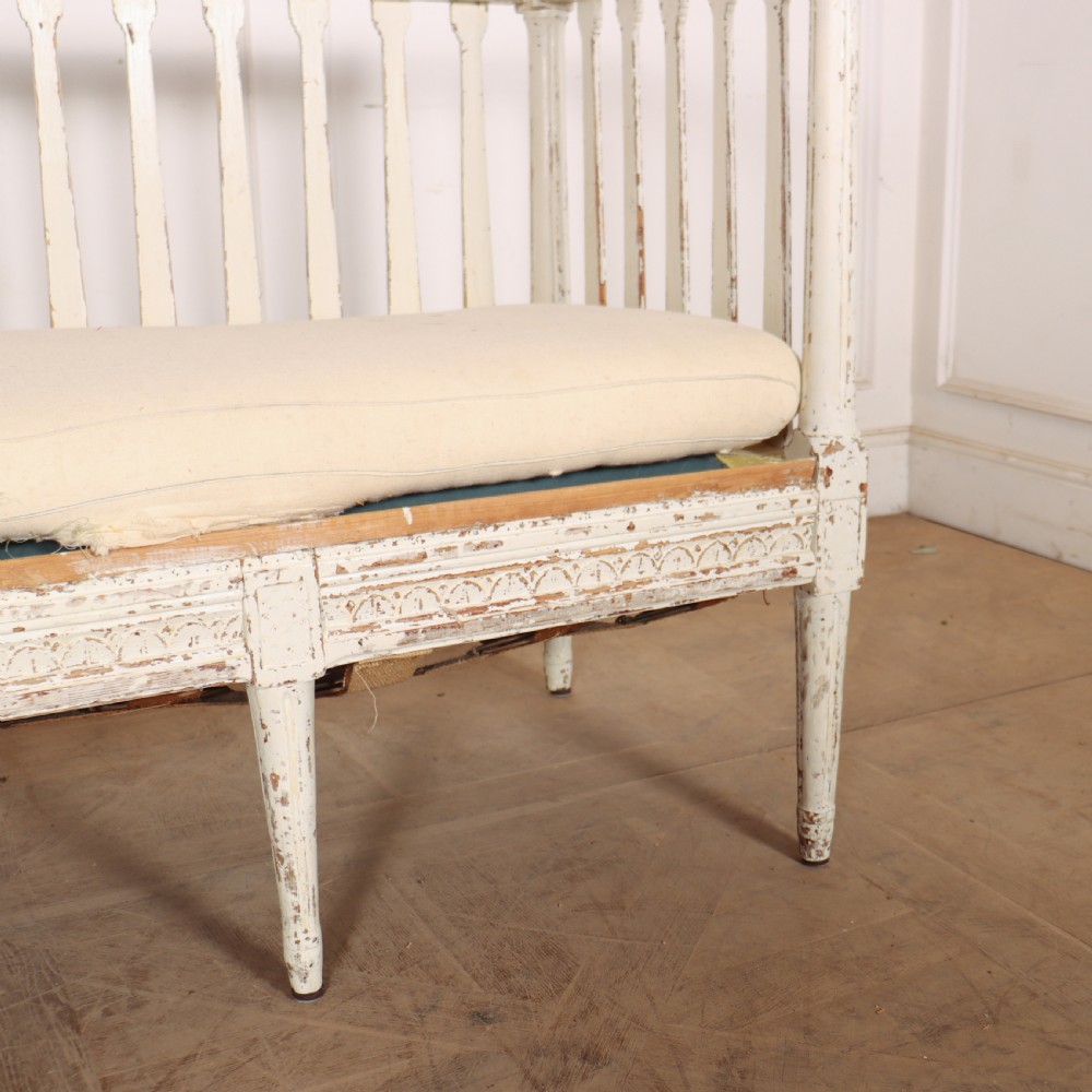 A long Gustavian style Swedish painted wooden settle bench featuring spindle sides and back, a distressed off-white finish, carved frame detailing, six tapered legs, and a fitted neutral upholstered cushion. View V