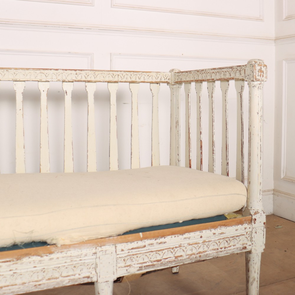 A long Gustavian style Swedish painted wooden settle bench featuring spindle sides and back, a distressed off-white finish, carved frame detailing, six tapered legs, and a fitted neutral upholstered cushion. View VI