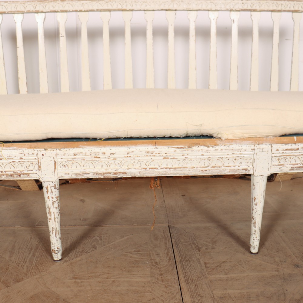 A long Gustavian style Swedish painted wooden settle bench featuring spindle sides and back, a distressed off-white finish, carved frame detailing, six tapered legs, and a fitted neutral upholstered cushion. View VII
