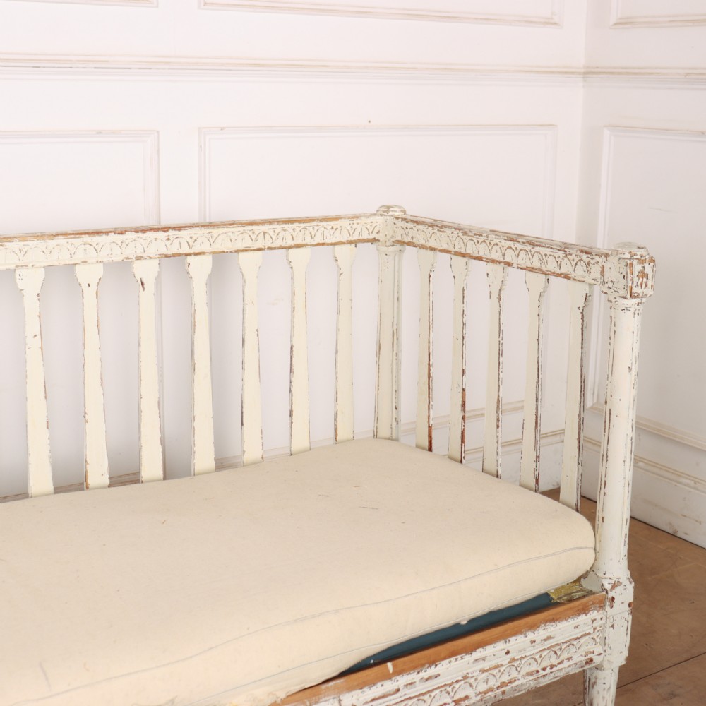A long Gustavian style Swedish painted wooden settle bench featuring spindle sides and back, a distressed off-white finish, carved frame detailing, six tapered legs, and a fitted neutral upholstered cushion. View VIII