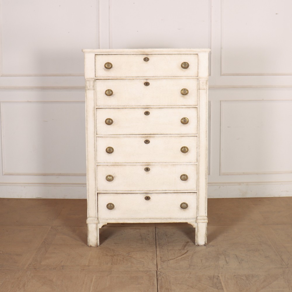 An off-white painted tallboy chest of drawers featuring six stacked drawers with round brass handles and keyhole escutcheons.