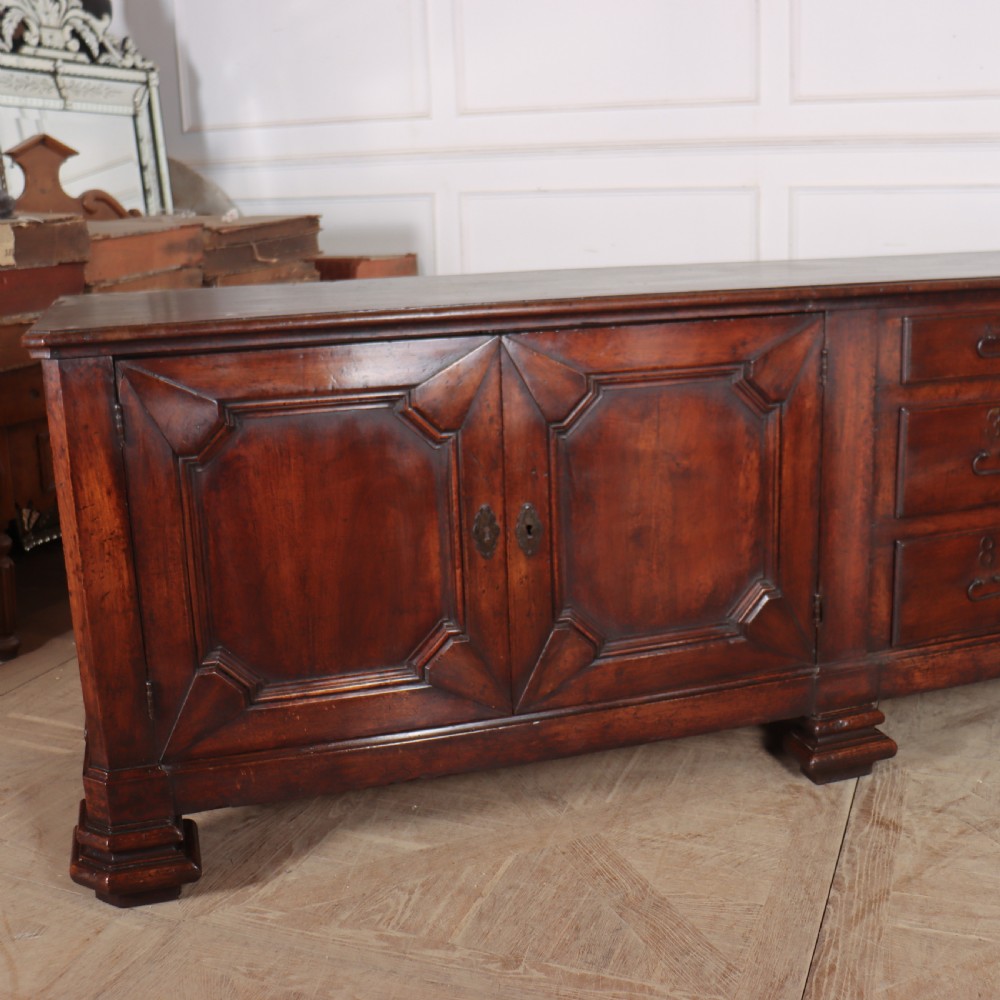 A long, monumental dark walnut Italian credenza featuring geometric moulded panel doors, a central column of drawers, and carved plinth feet. View III