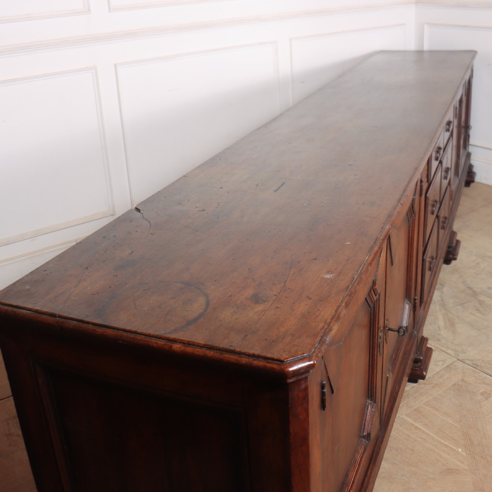 A long, monumental dark walnut Italian credenza featuring geometric moulded panel doors, a central column of drawers, and carved plinth feet. View XIII