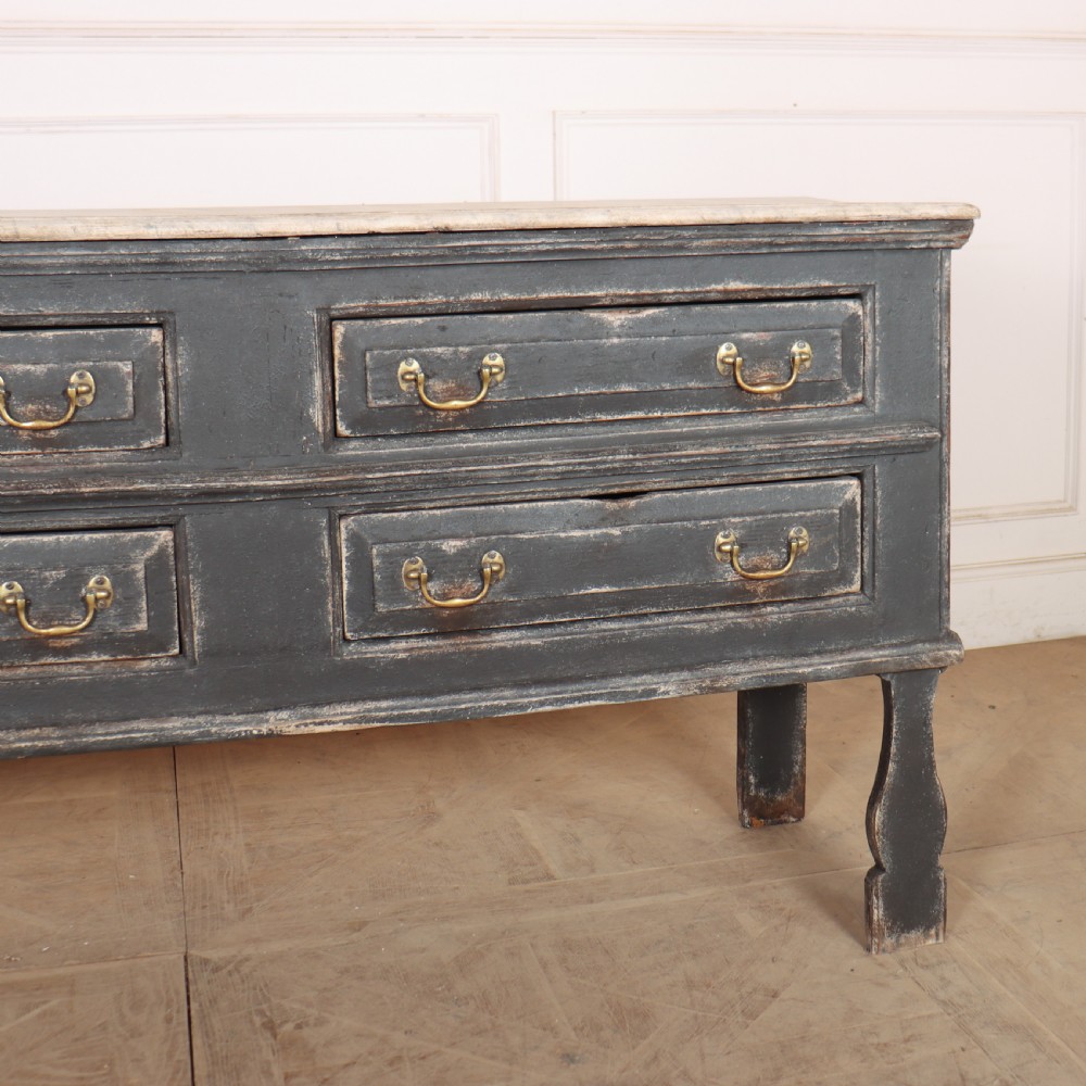An English painted dresser base featuring a dark charcoal finish, four wide drawers with brass drop handles, shaped corner legs, and a light marble top. View III