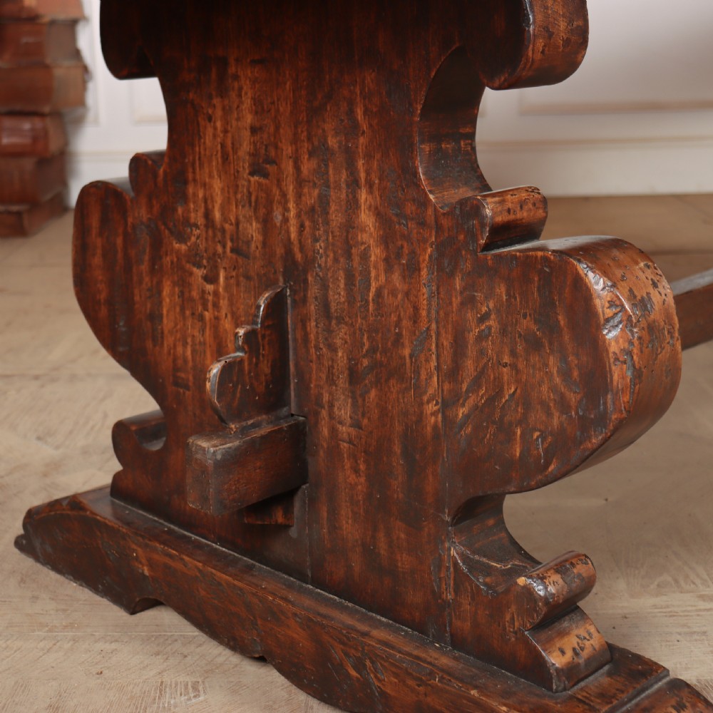 A long antique dark walnut refectory dining table featuring a thick plank top resting on carved trestle supports joined by a central stretcher. View III