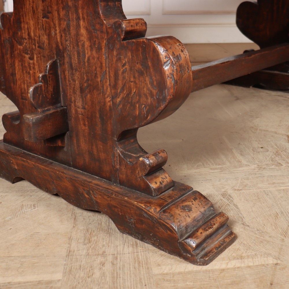 A long antique dark walnut refectory dining table featuring a thick plank top resting on carved trestle supports joined by a central stretcher. View IV