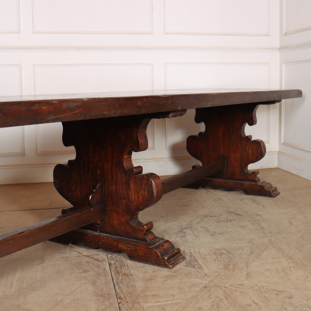 A long antique dark walnut refectory dining table featuring a thick plank top resting on carved trestle supports joined by a central stretcher. View VI