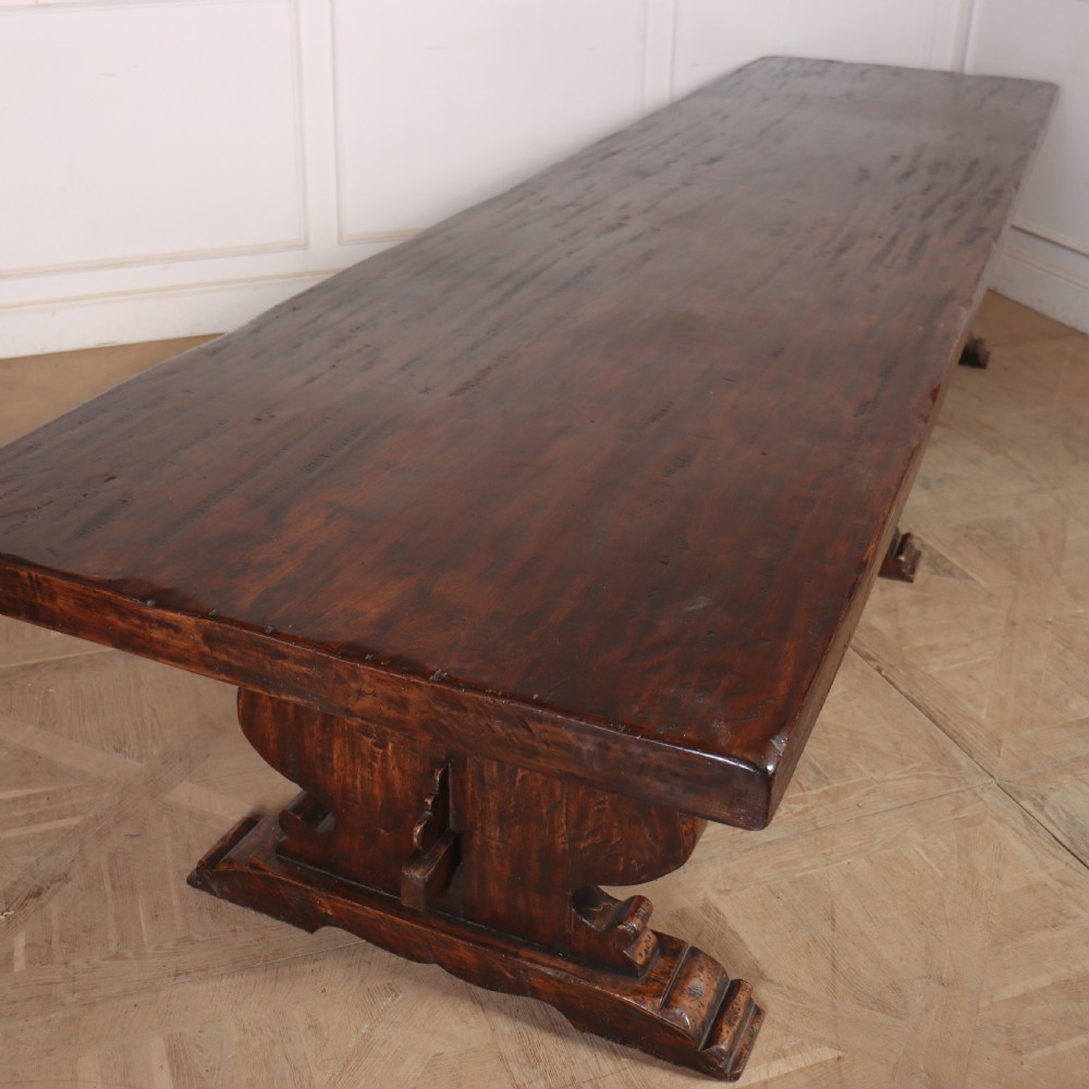 A long antique dark walnut refectory dining table featuring a thick plank top resting on carved trestle supports joined by a central stretcher. View VII