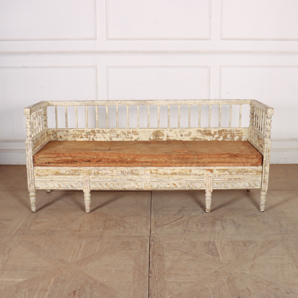 An antique Swedish wooden settle bench featuring a distressed off-white painted finish, vertical spindle backrest, carved apron, fluted legs, and a padded seat cushion.