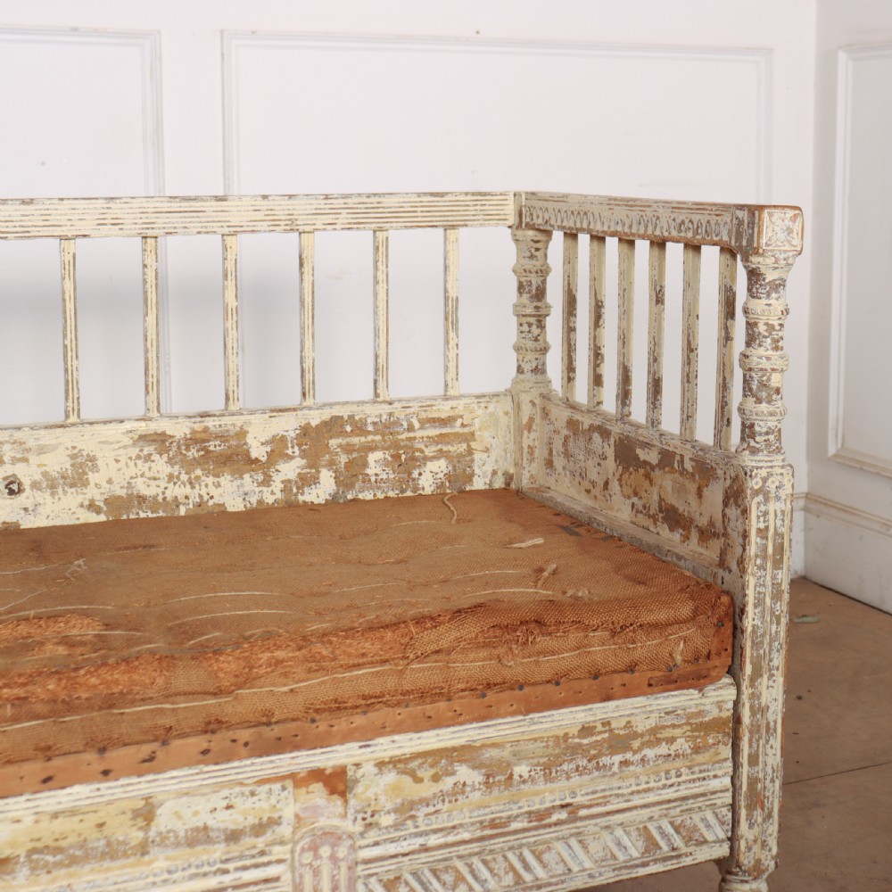 An antique Swedish wooden settle bench featuring a distressed off-white painted finish, vertical spindle backrest, carved apron, fluted legs, and a padded seat cushion. View II