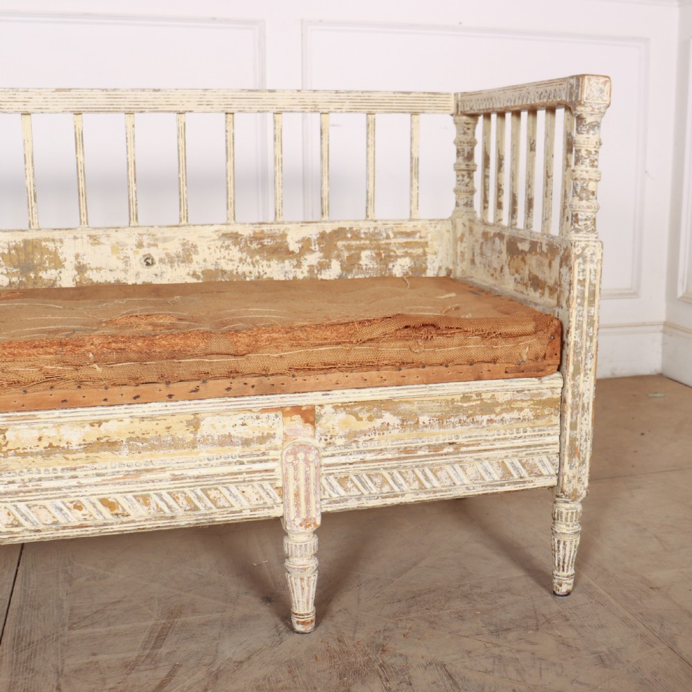 An antique Swedish wooden settle bench featuring a distressed off-white painted finish, vertical spindle backrest, carved apron, fluted legs, and a padded seat cushion. View III