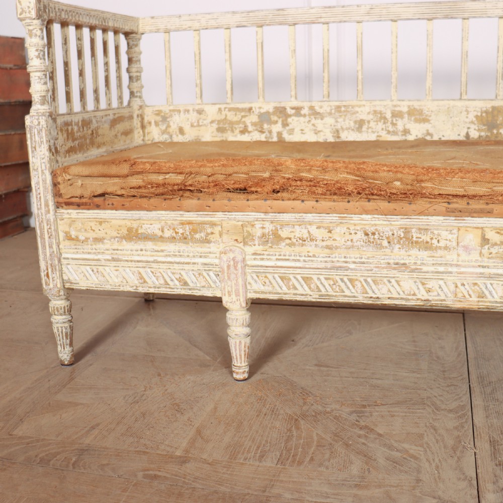 An antique Swedish wooden settle bench featuring a distressed off-white painted finish, vertical spindle backrest, carved apron, fluted legs, and a padded seat cushion. View V