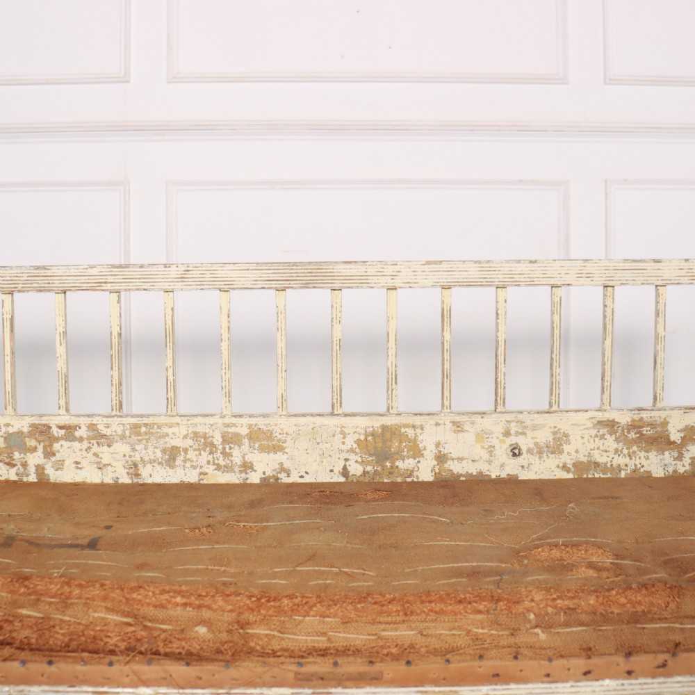 An antique Swedish wooden settle bench featuring a distressed off-white painted finish, vertical spindle backrest, carved apron, fluted legs, and a padded seat cushion. View VI