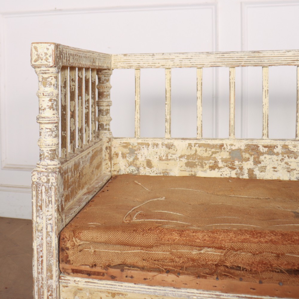 An antique Swedish wooden settle bench featuring a distressed off-white painted finish, vertical spindle backrest, carved apron, fluted legs, and a padded seat cushion. View VII