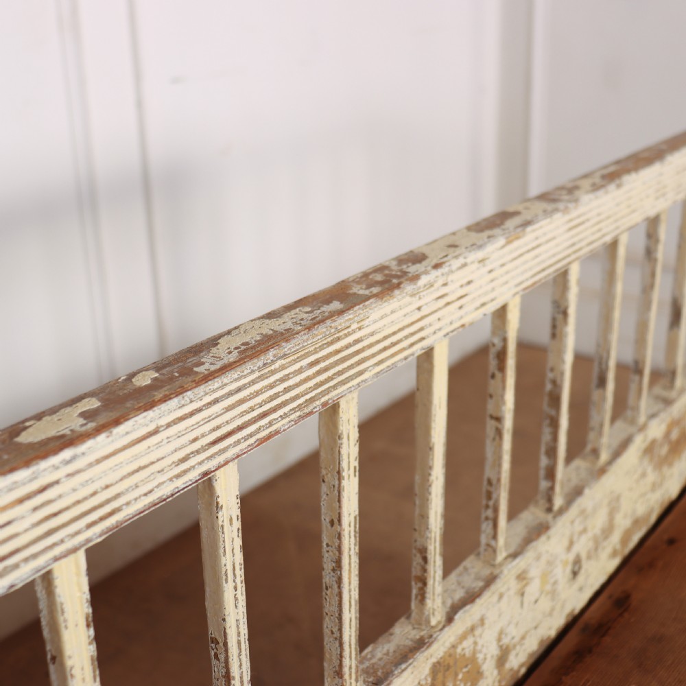 An antique Swedish wooden settle bench featuring a distressed off-white painted finish, vertical spindle backrest, carved apron, fluted legs, and a padded seat cushion. View VIII