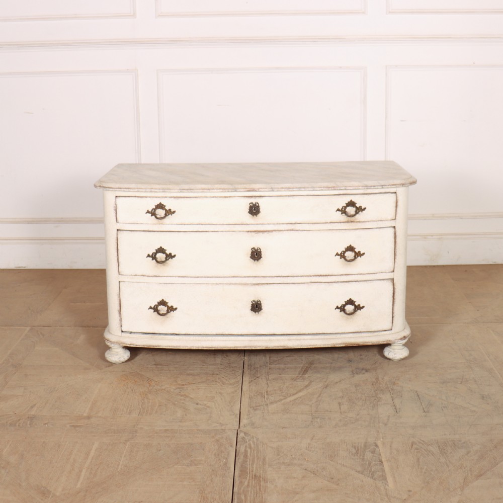 An off-white painted French bow-front commode with three wide drawers, dark metal drop handles, and a light grey marble top.