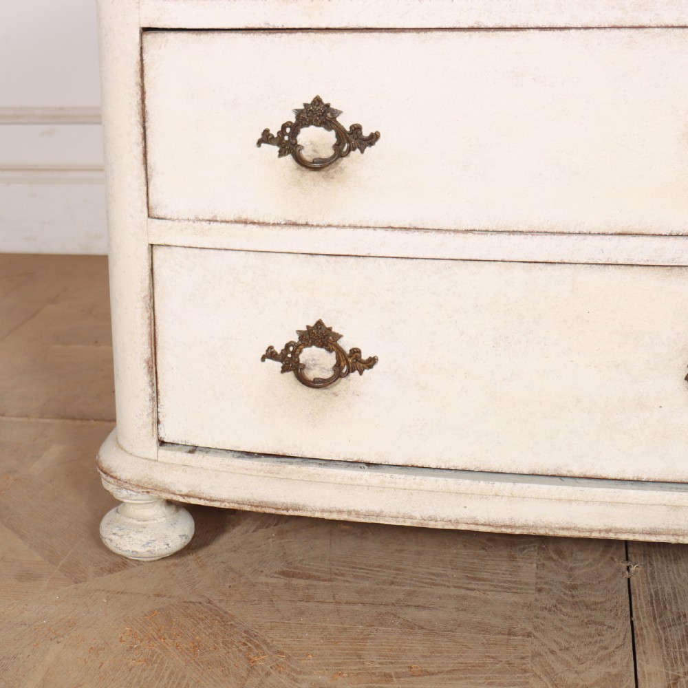 An off-white painted French bow-front commode with three wide drawers, dark metal drop handles, and a light grey marble top. View V