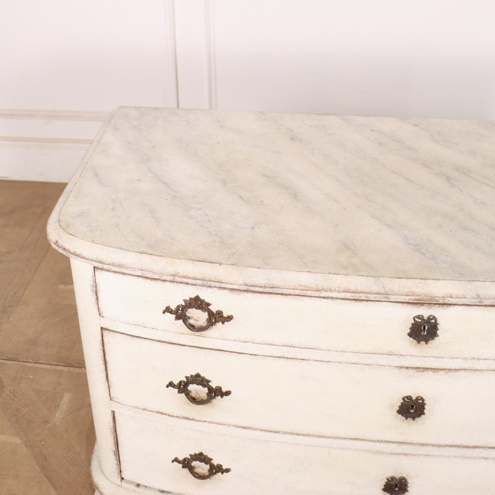 An off-white painted French bow-front commode with three wide drawers, dark metal drop handles, and a light grey marble top. View IX