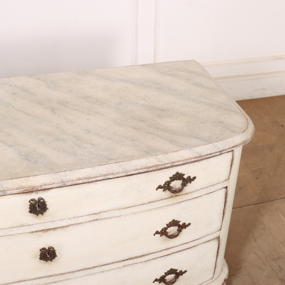 An off-white painted French bow-front commode with three wide drawers, dark metal drop handles, and a light grey marble top. View X