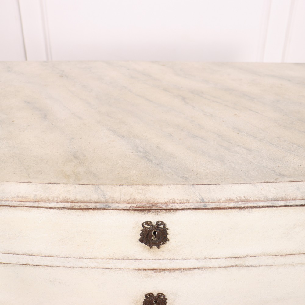 An off-white painted French bow-front commode with three wide drawers, dark metal drop handles, and a light grey marble top. View XI