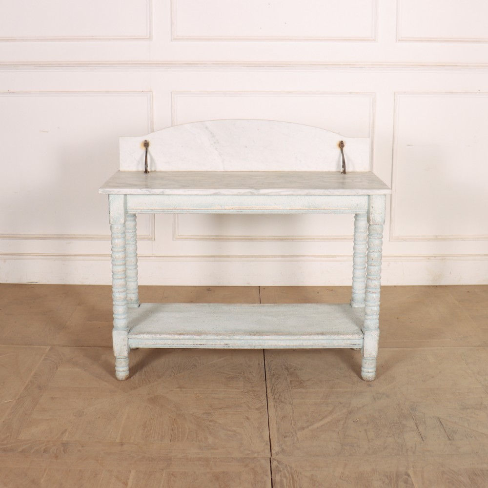 An antique painted wooden washstand featuring bobbin turned legs, a lower shelf, a pale blue distressed finish, and a white marble top with a splashback.