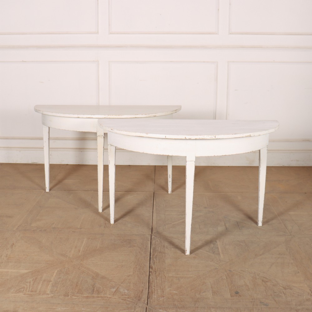 A pair of Swedish demi-lune console tables featuring a distressed off-white painted finish and slender tapered legs.