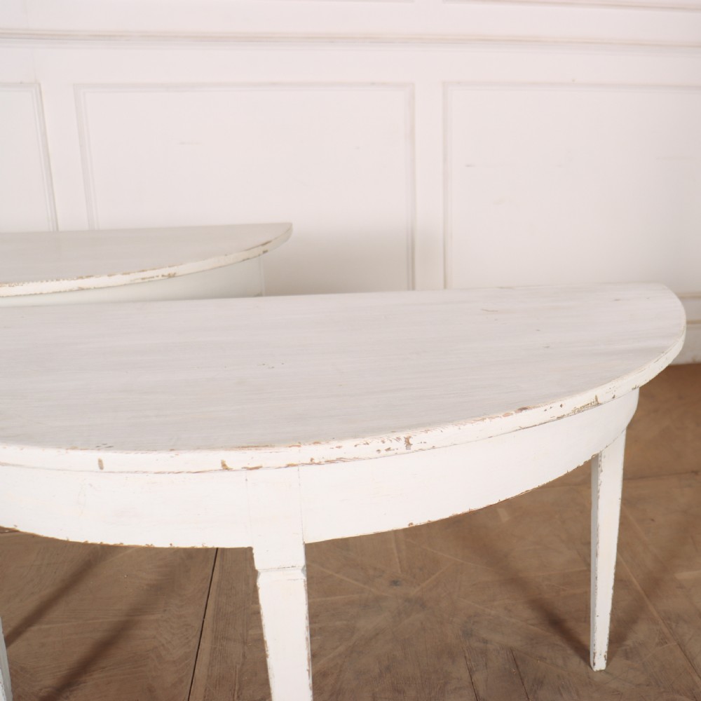A pair of Swedish demi-lune console tables featuring a distressed off-white painted finish and slender tapered legs. View V