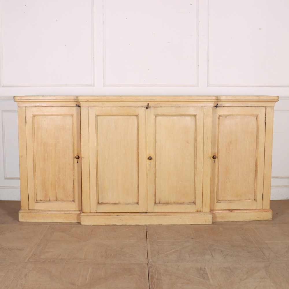 A wide English pine breakfront cupboard featuring four recessed panel doors, brass knobs, and a projecting central section.