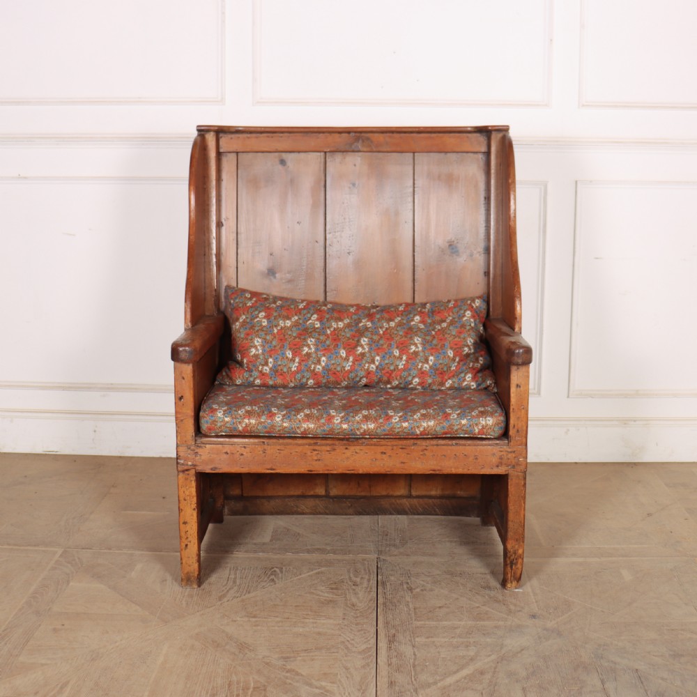 A high-backed antique pine wooden settle featuring tall vertical plank panels, curved side wings, and a floral upholstered seat cushion.