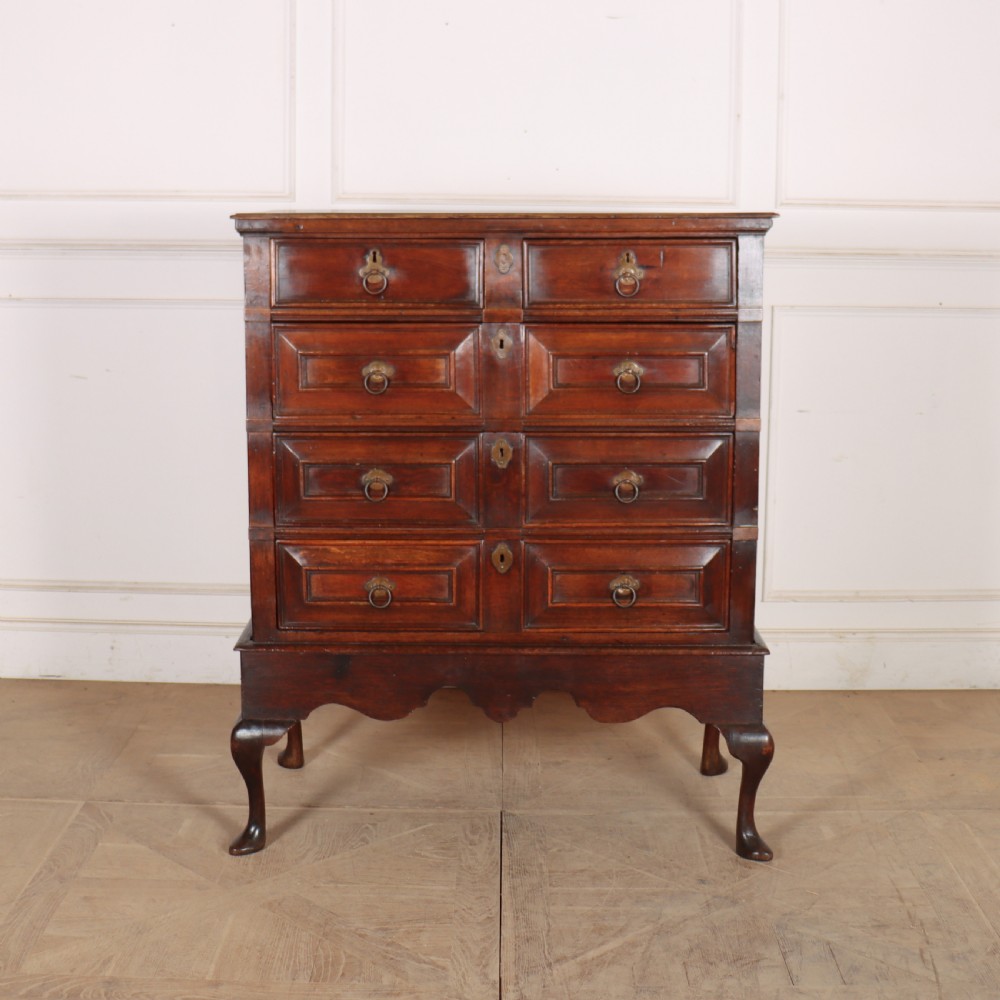 A warm brown wooden antique chest on stand with geometric moulded drawer fronts, brass drop handles, and slender cabriole legs.