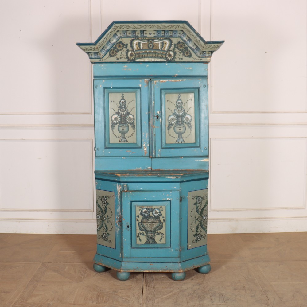 A tall two-tier Gustavian painted pine cabinet featuring a distressed blue finish, hand-painted urn panels, a shaped pediment with date inscription, and turned bun feet.