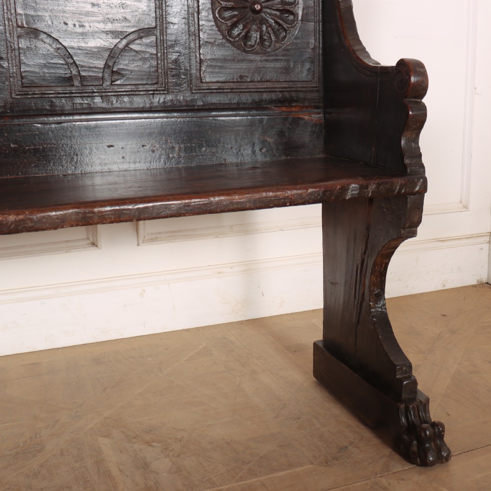 A dark brown carved walnut bench with a panelled backrest featuring carved rosette medallions and detailed carved feet. View V