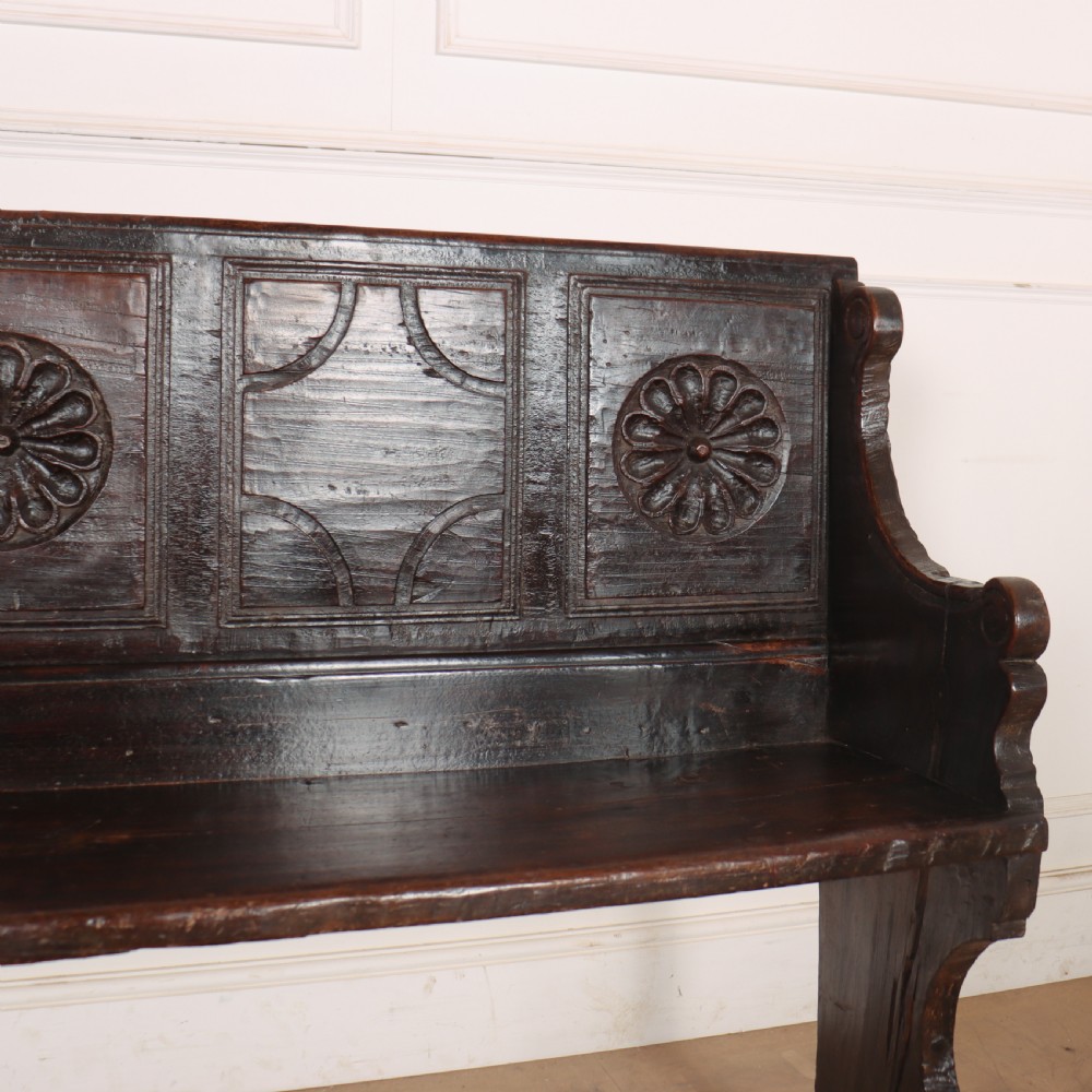 A dark brown carved walnut bench with a panelled backrest featuring carved rosette medallions and detailed carved feet. View VI