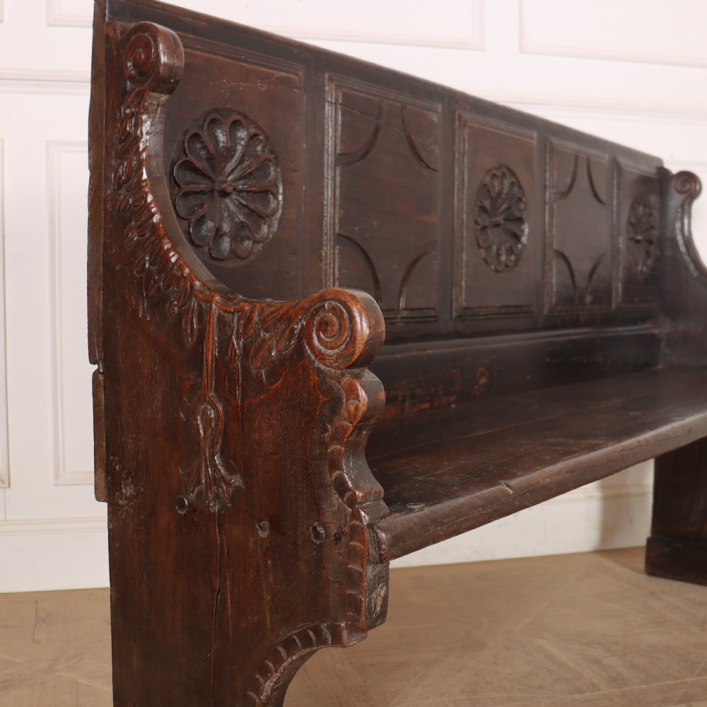 A dark brown carved walnut bench with a panelled backrest featuring carved rosette medallions and detailed carved feet. View IX